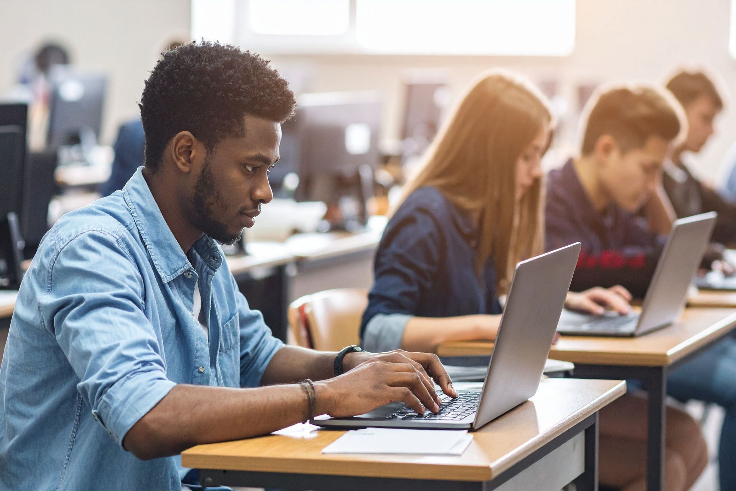 Student working on a laptop in a classroom while other students work in the background.